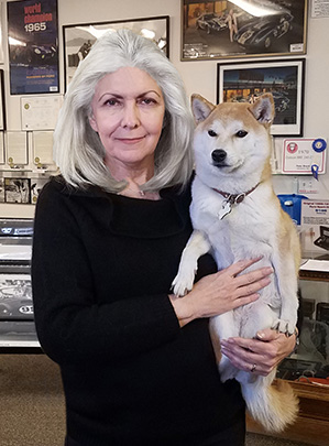 Gayle Brock poses for a photo with her shiba inu. Behind them is a wall with framed movie posters and a certificate.